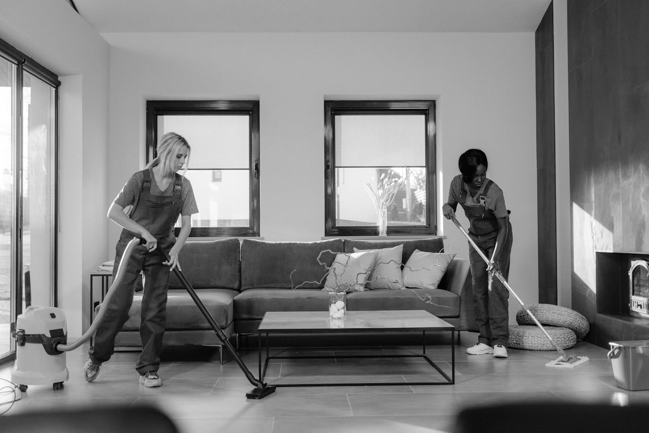 home-services-02 Two women in work clothes cleaning a modern living room using a vacuum and mop.